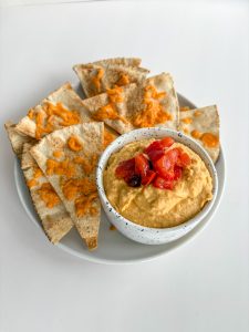 Cheese-covered pita on a plate beside a bowl of hummus and red peppers.