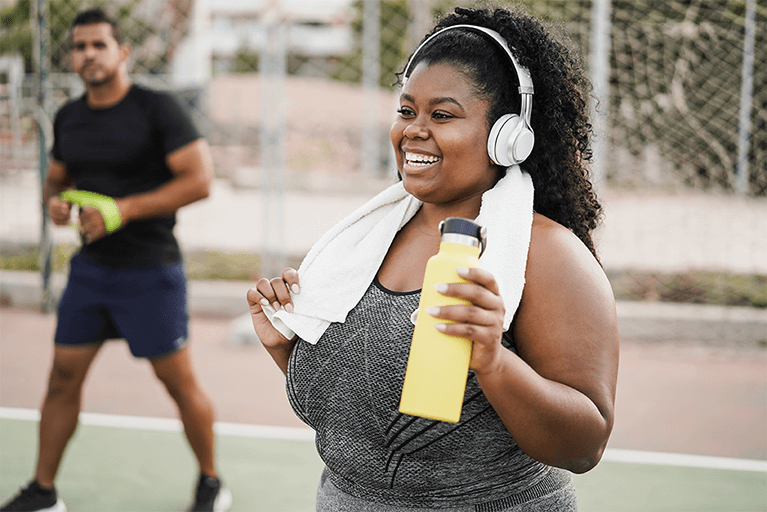 Une femme souriante en tenue de sport qui tient une bouteille d’eau et une serviette.