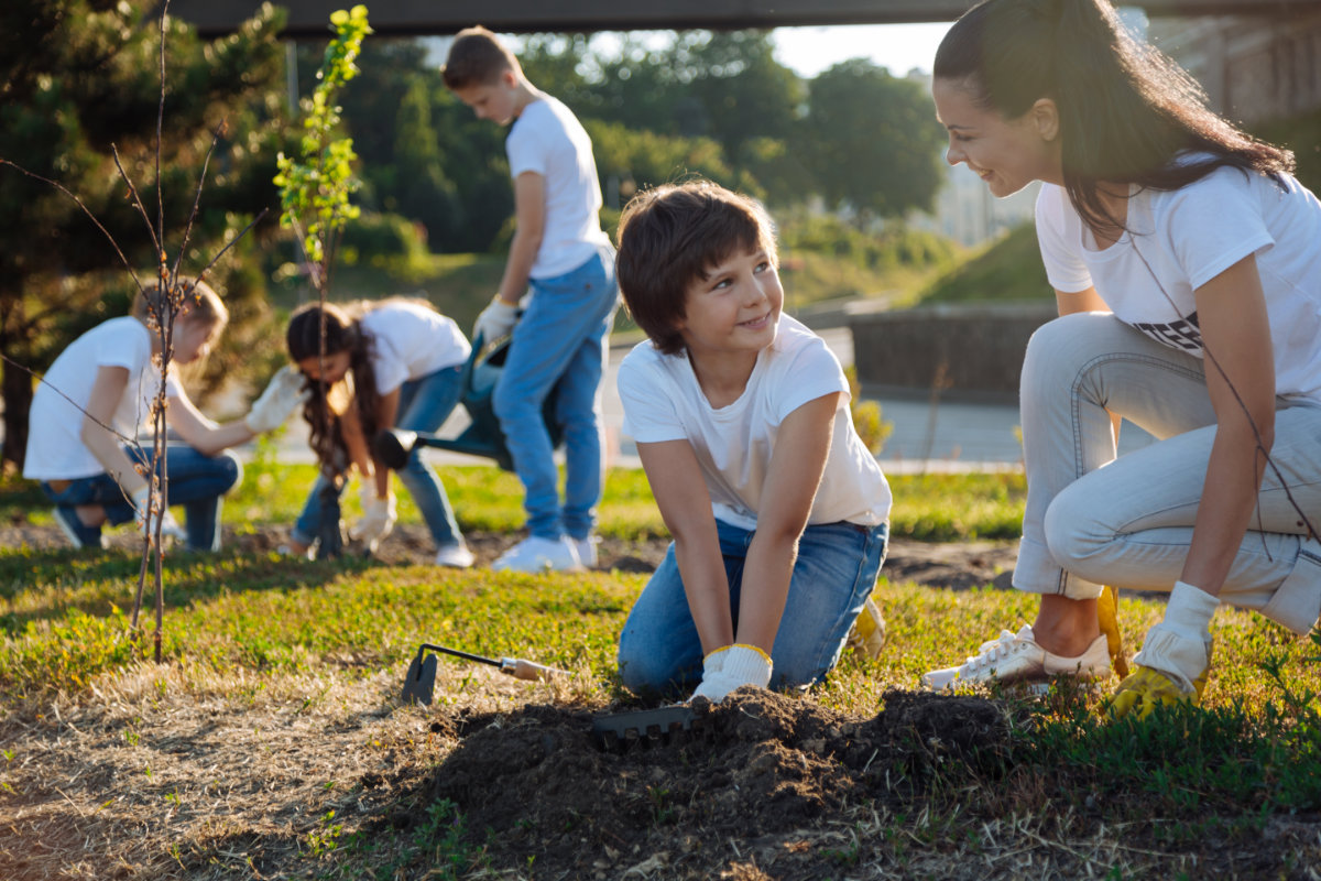 A kneeling woman teaching a young boy how to garden while more youth are standing in the background also gardening