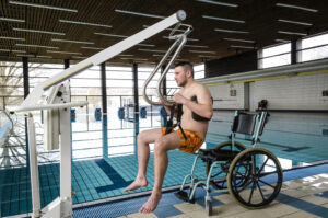 Un appareil soulève un homme de son fauteuil roulant pour le faire entrer dans une piscine couverte. Un appareil soulève un homme de son fauteuil roulant pour le faire entrer dans une piscine couverte.