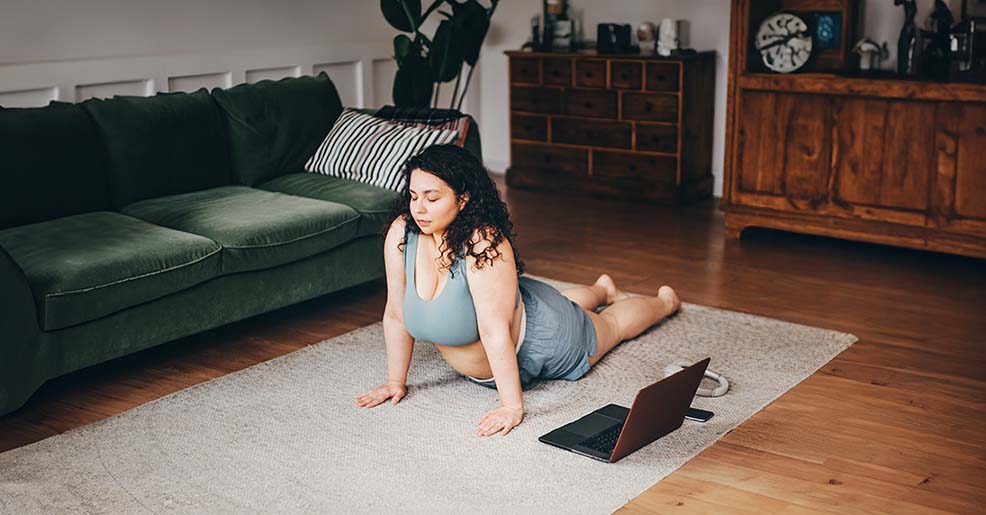 Une femme fait du yoga à côté d'un ordinateur portable sur le sol d'un salon.