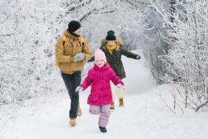 A man, woman and child running on a snow-covered nature trail surrounded by trees to beat the winter blues.