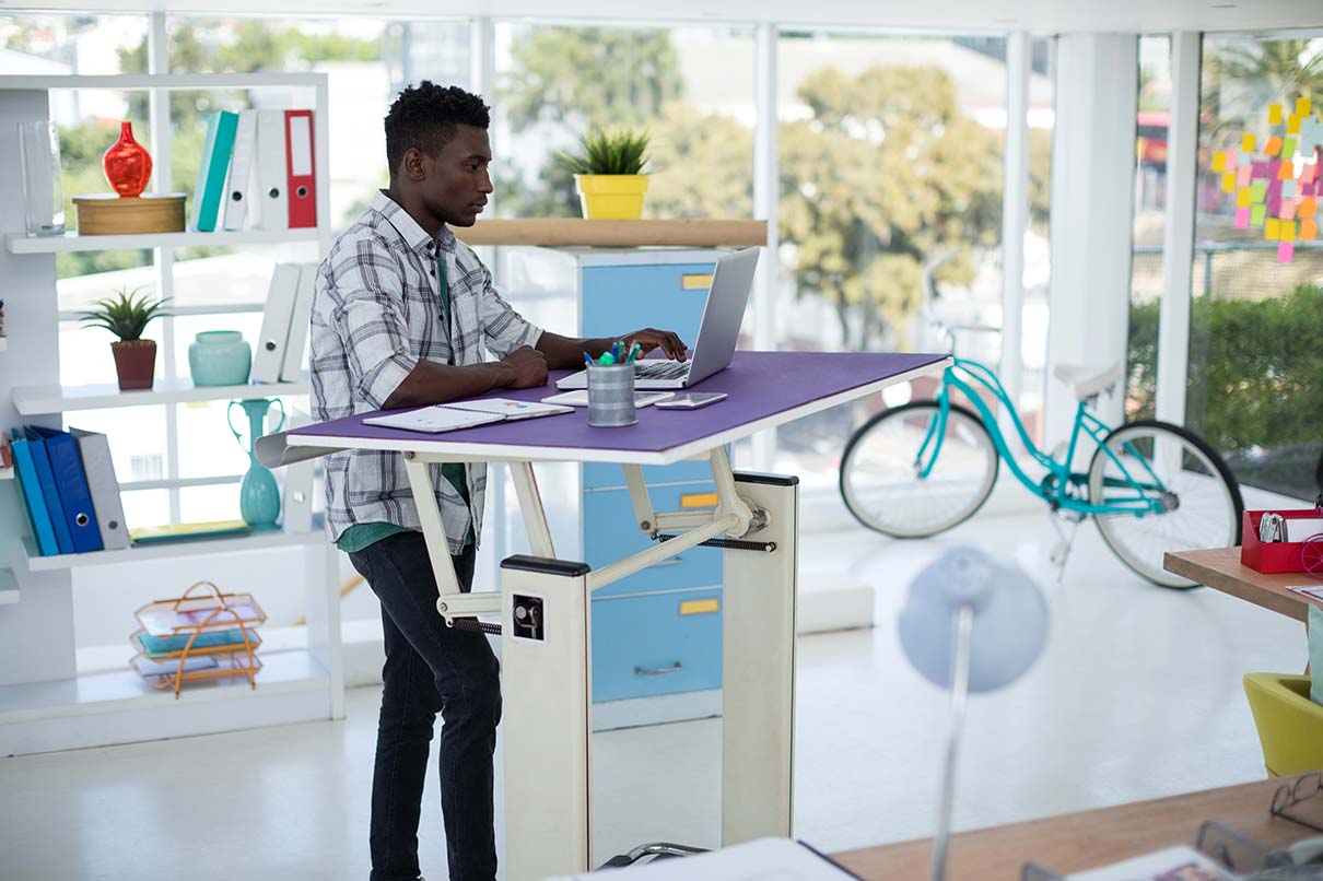 A man using a laptop on an adjustable standing desk. A man using a laptop on an adjustable standing desk.