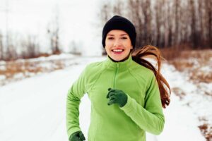 Une femme souriante faisant du jogging sur un sentier naturel enneigé en décembre.