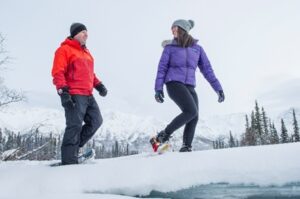 Two people snowshoeing on a nature trail with snow-covered mountains in the background