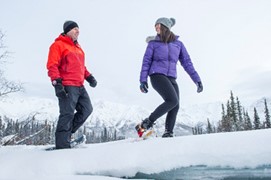 Deux personnes faisant de la raquette sur un sentier naturel avec des montagnes enneigées en arrière-plan.