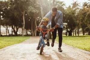    A man helping his son ride a bike on a park path surrounded by trees.