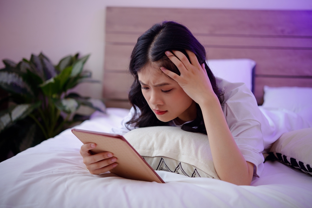 A worried-looking woman lying in bed while looking at a tablet. A worried-looking woman lying in bed while looking at a tablet.