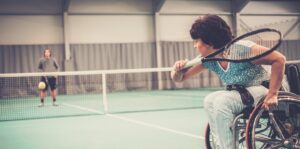 A woman playing wheelchair tennis indoors against a man standing on the other side of the net.