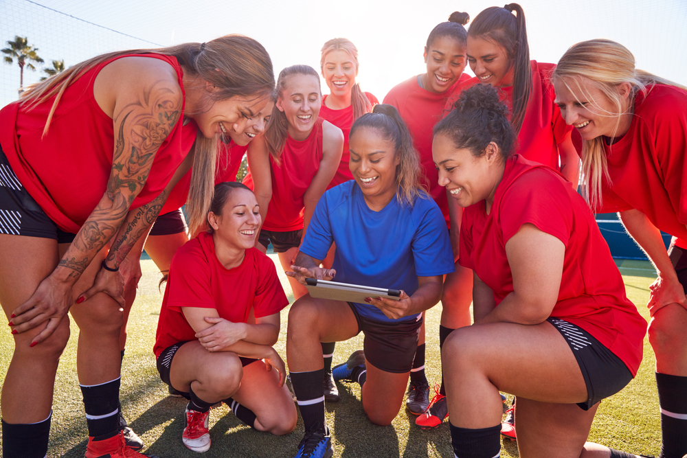 A group of women in red shirts looking at a tablet on a sports field. A group of women in red shirts looking at a tablet on a sports field.