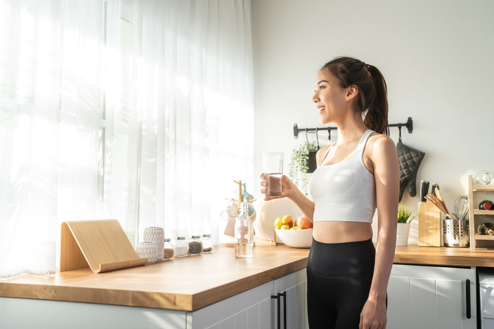 Une femme souriante en tenue de sport tenant un verre d’eau dans une cuisine. 