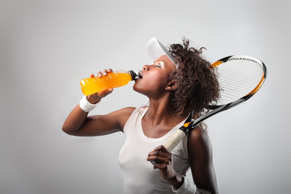 Une femme buvant une boisson pour sportifs tout en tenant une raquette de tennis. 