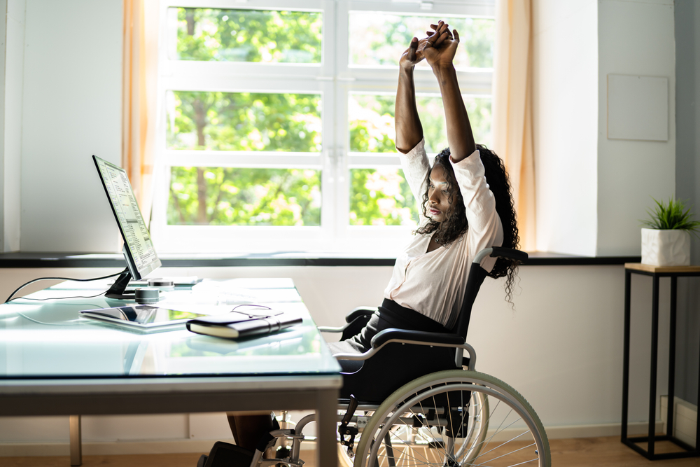 Une femme en fauteuil roulant étire ses bras au-dessus de la tête devant un bureau