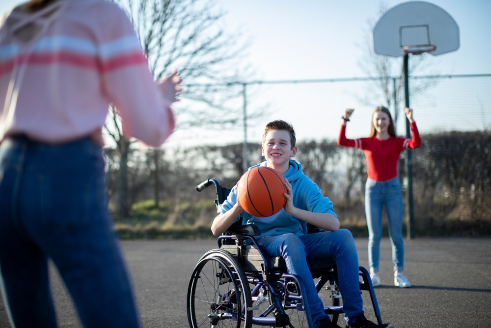Un garçon dans un fauteuil roulant joue au basketball avec deux filles à l’extérieur. Un garçon dans un fauteuil roulant joue au basketball avec deux filles à l’extérieur.