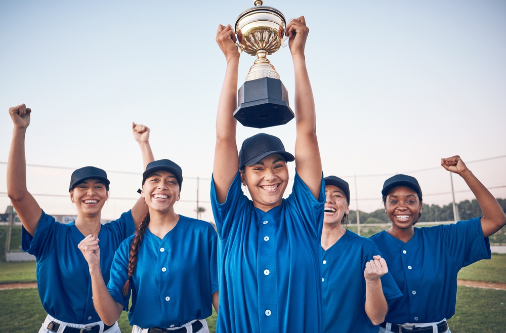 Une joueuse de baseball tient un trophée pendant que ses coéquipières célèbrent derrière elle. Une joueuse de baseball tient un trophée pendant que ses coéquipières célèbrent derrière elle.