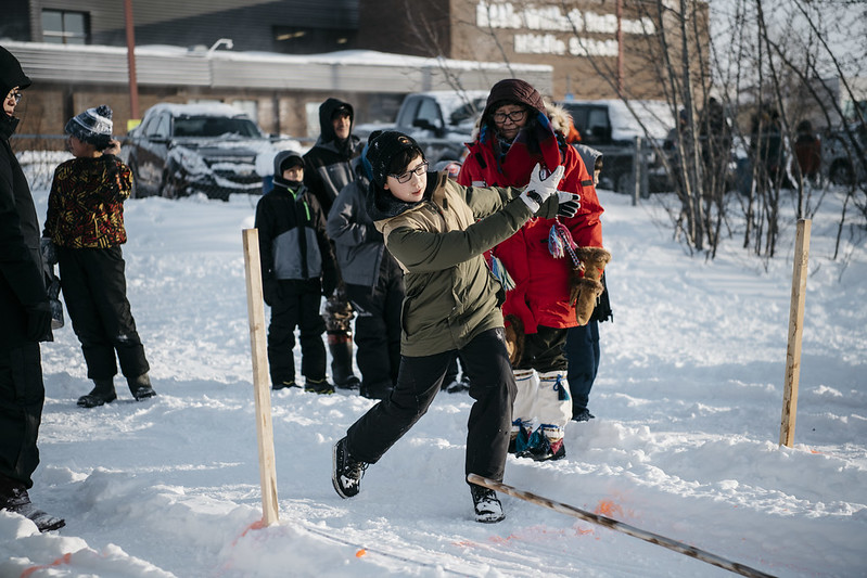 A group of kids playing the Dene game called snow snake in a snow-covered field. A group of kids playing the Dene game called snow snake in a snow-covered field.