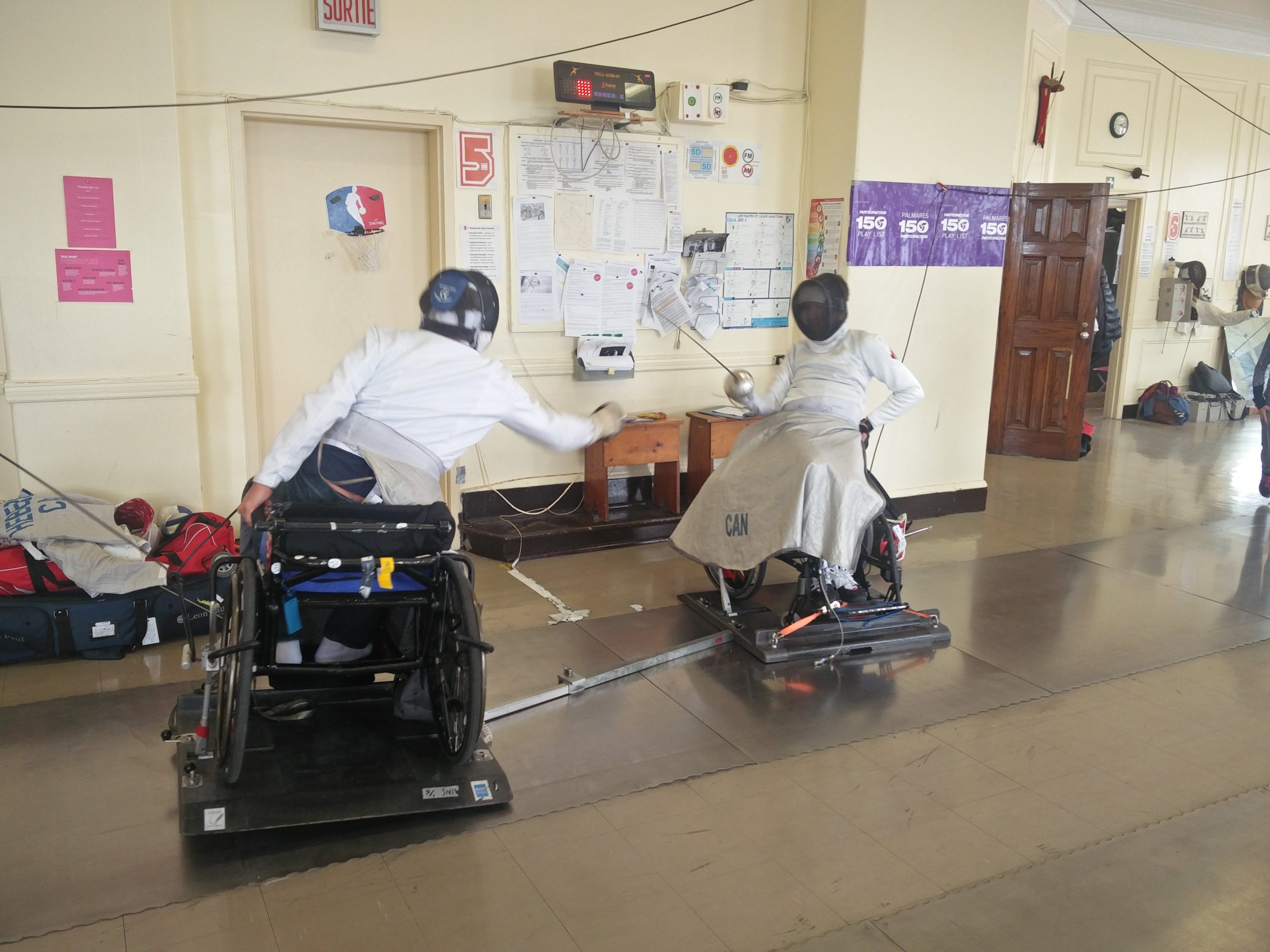 Deux personnes en fauteuils roulants font de l’escrime dans une salle de classe. Deux personnes en fauteuils roulants font de l’escrime dans une salle de classe.