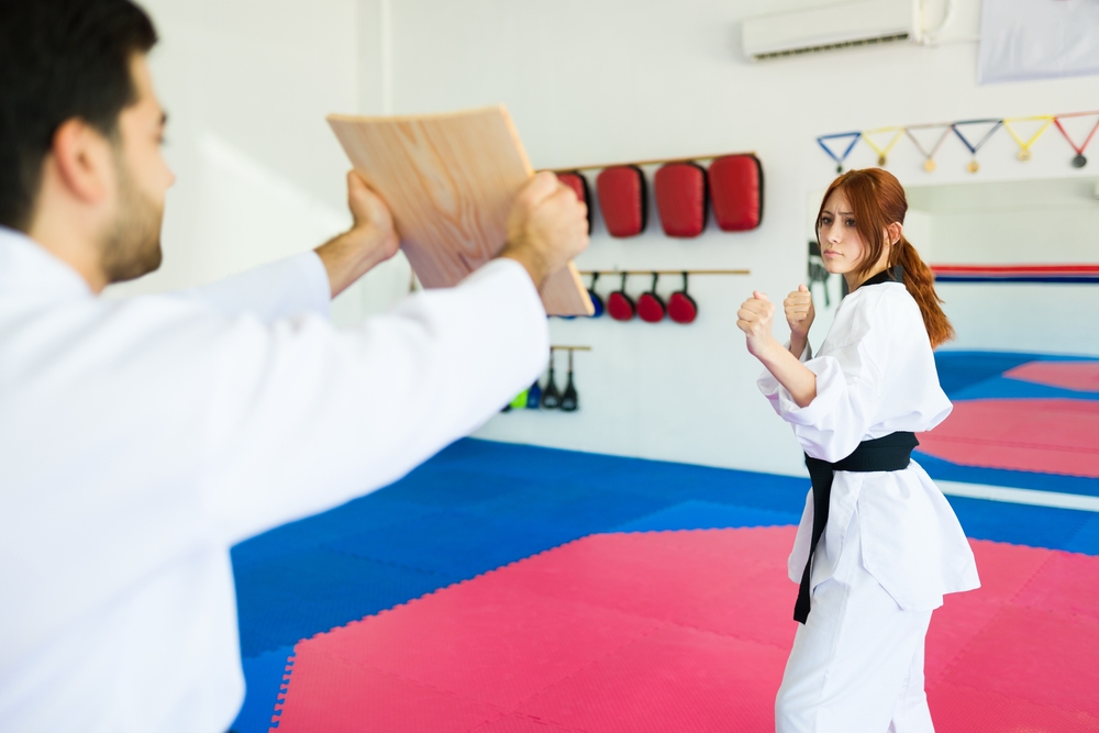 Une femme en tenue de taekwondo s’apprête à casser une planche de bois tenue par un homme. Une femme en tenue de taekwondo s’apprête à casser une planche de bois tenue par un homme.
