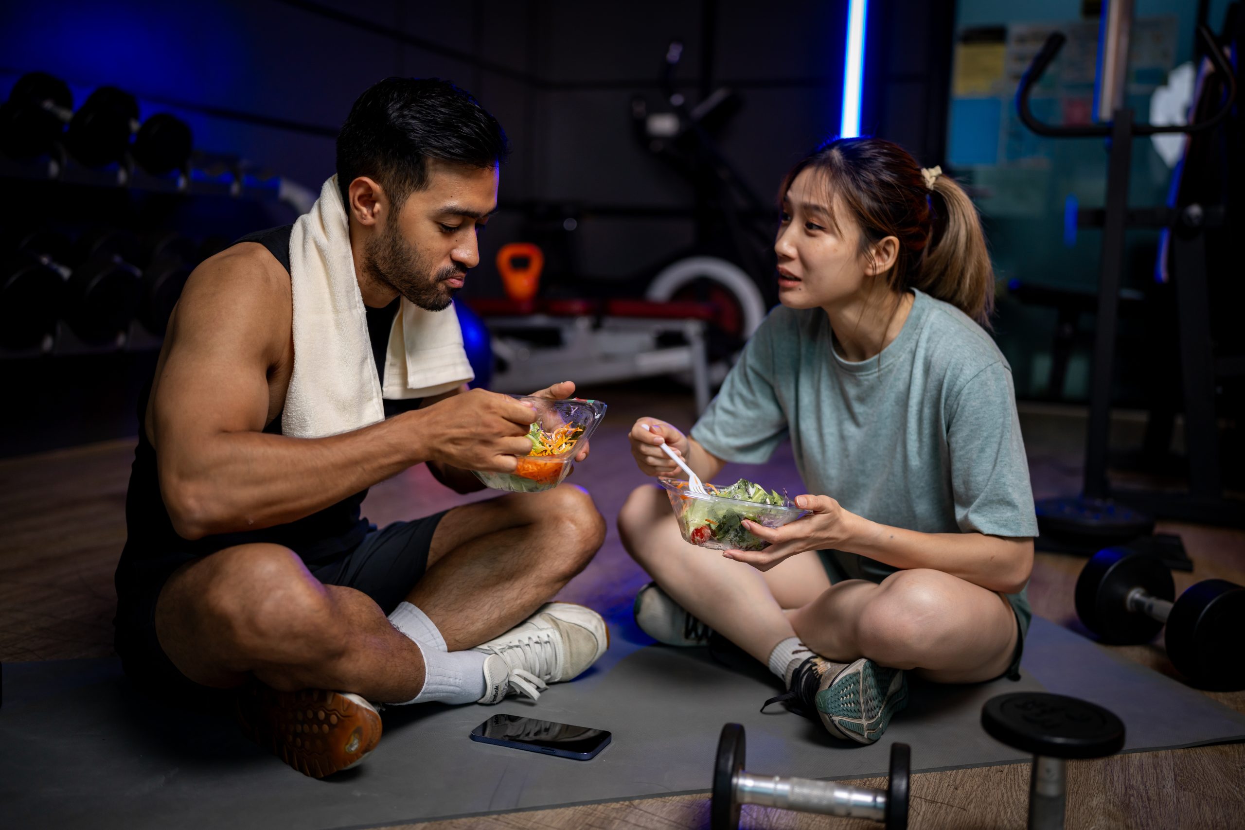 A man and woman eating salads on the floor of a gym. A man and woman eating salads on the floor of a gym.