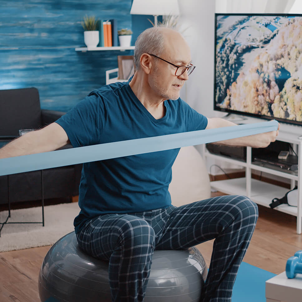 Un homme étire une bande élastique avec ses bras assis sur un ballon d’équilibre placé sur le plancher du salon.