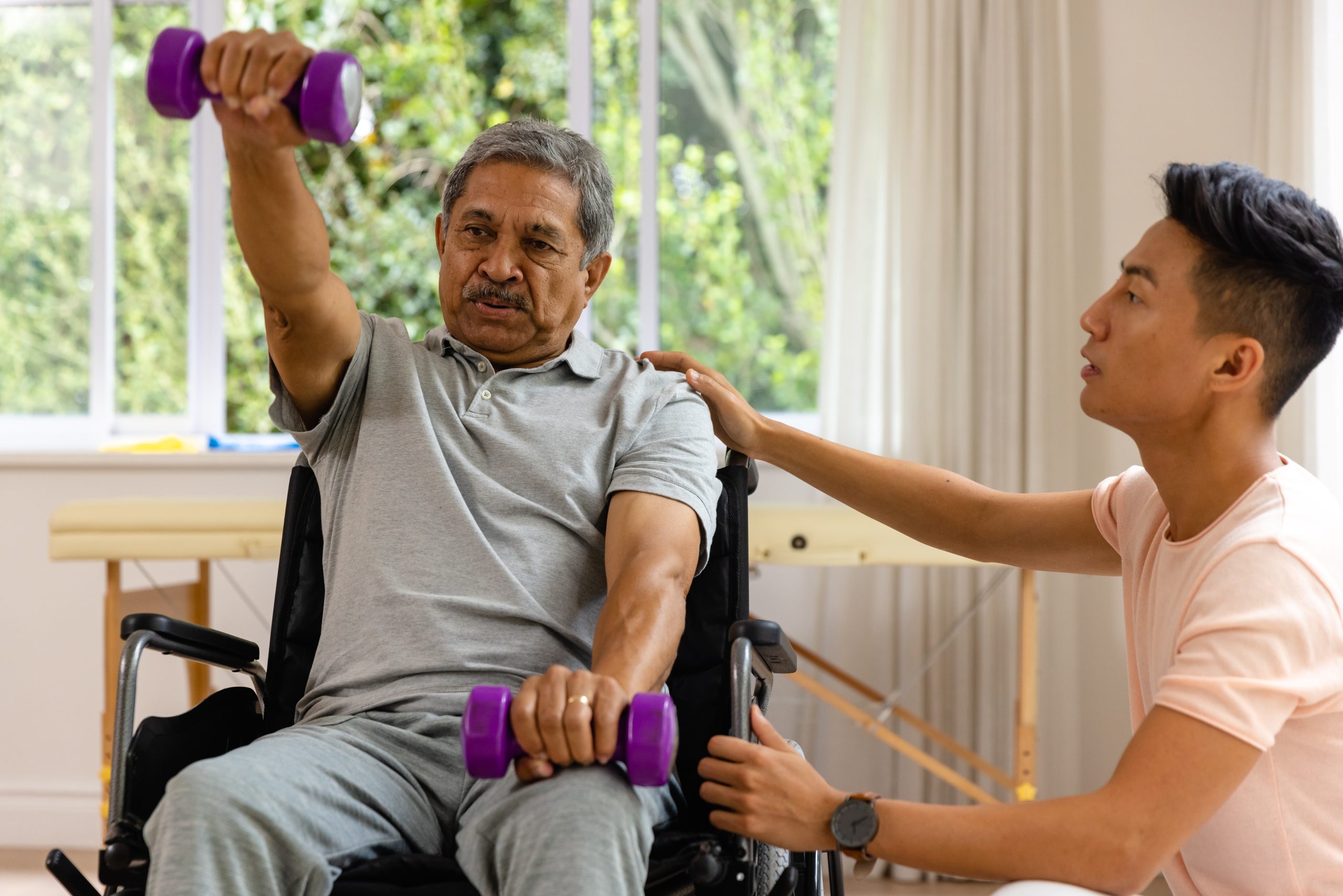 Un jeune homme aide un homme plus âgé assis dans un fauteuil roulant à faire des exercices avec des haltères. Un jeune homme aide un homme plus âgé assis dans un fauteuil roulant à faire des exercices avec des haltères.