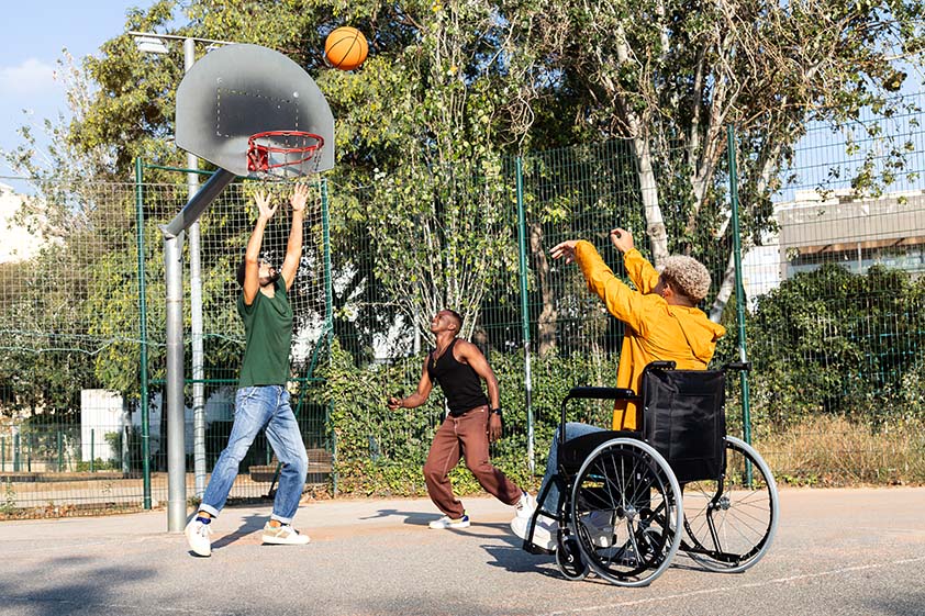 A man in a wheelchair playing basketball with two other men on an outdoor court. A man in a wheelchair playing basketball with two other men on an outdoor court.