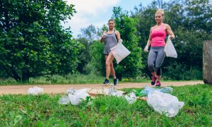 Two women jogging on a nature trail while carrying plastic bags.