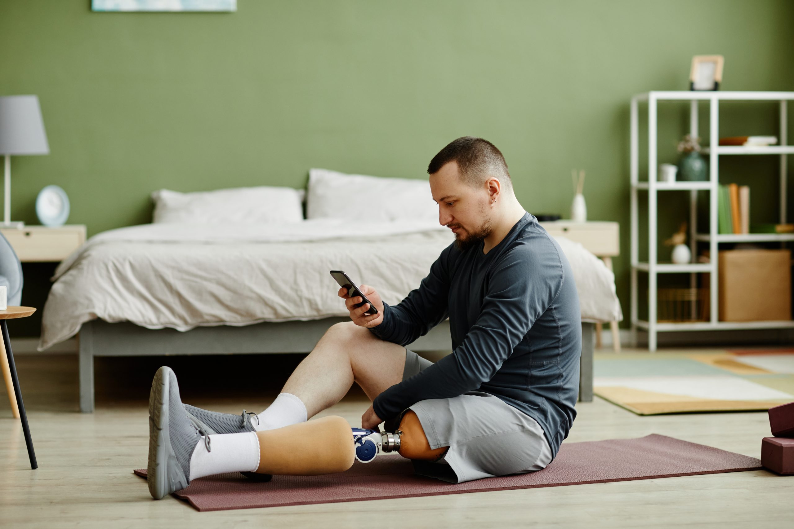 A man checking his phone while sitting on a yoga mat on a bedroom floor.